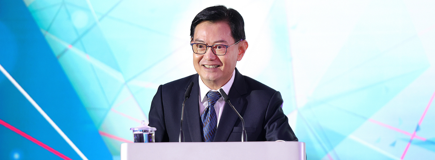 Lawrence Wong at podium with microphones, wearing suit and glasses, smiling. Blue geometric background.